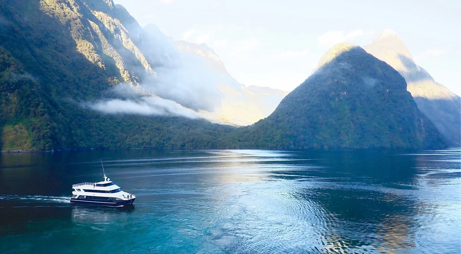 Small cruise boat dwarfed by Mitre Peak and the cloud-wreathed cliffs of Milford Sound