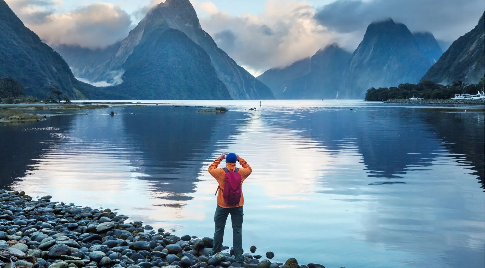 A hiker looking out over Milford Sound's mountains at sunrise from the rocky shoreline